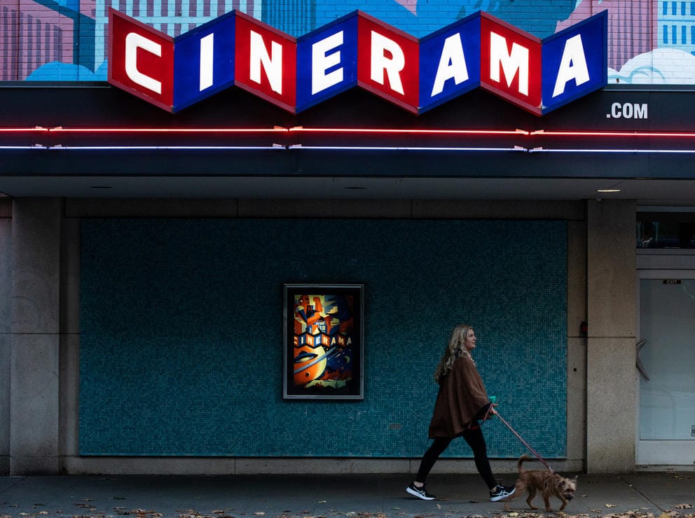 Woman walks her dog in front of a dark facade, up top red and blue squares with white letters spell out CINERAMA