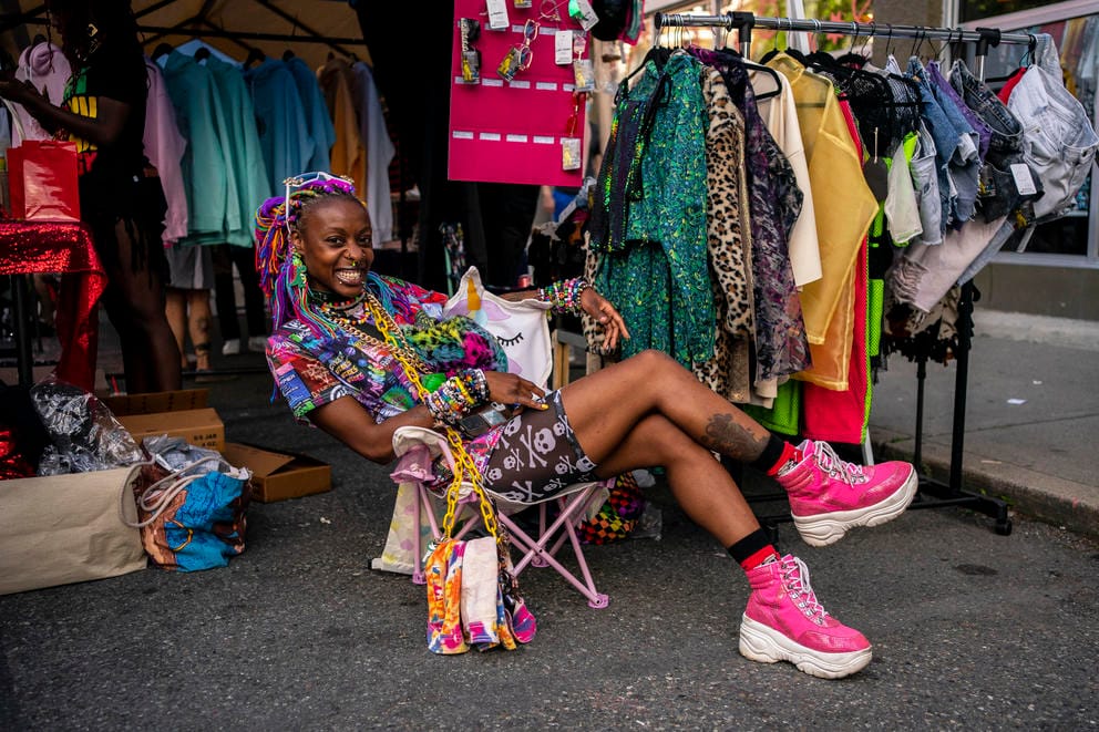 Andreya Taylor reclines in a camp chair wearing pink shoes colorful clothing. A rack of more colorful is behind her. 