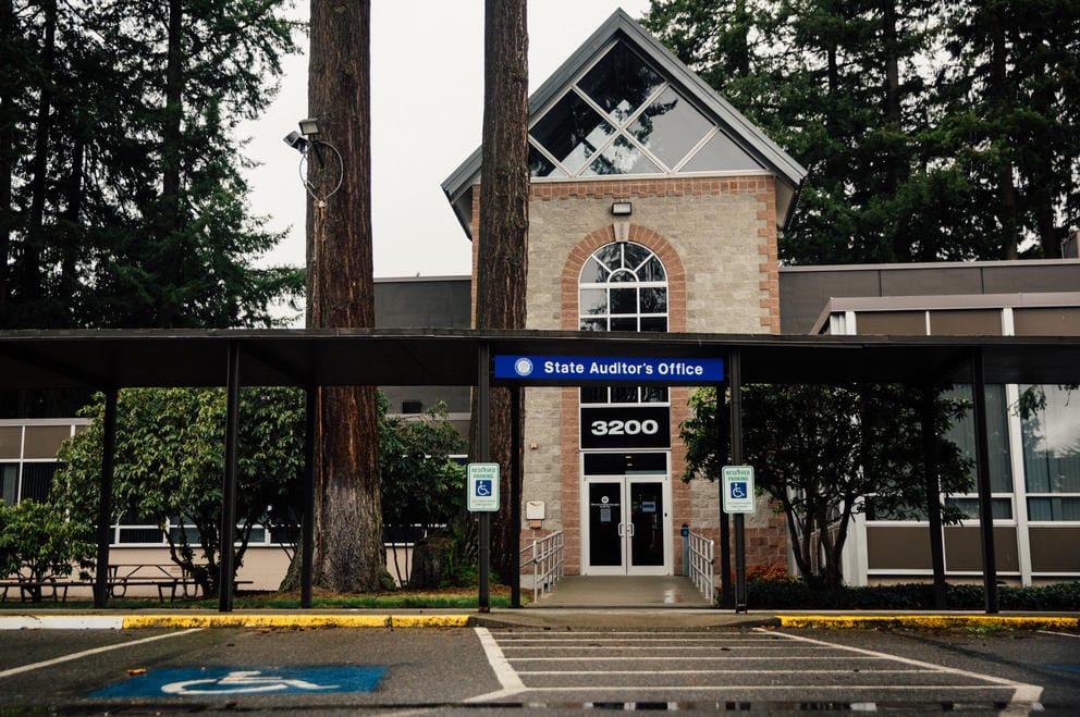 A view of the outside of the State Auditor's Office with tall evergreen trees in the background. A blue sign in front reads 