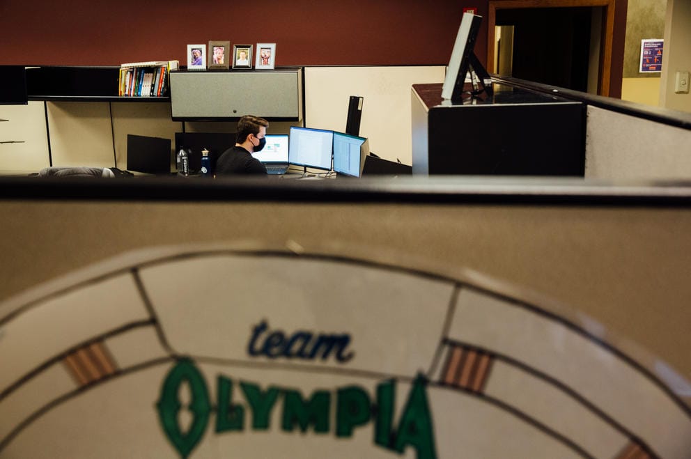Dylan Wharton, an assistant auditor, faces away as he works at his computer in his cubicle. A sign in the foreground reads 