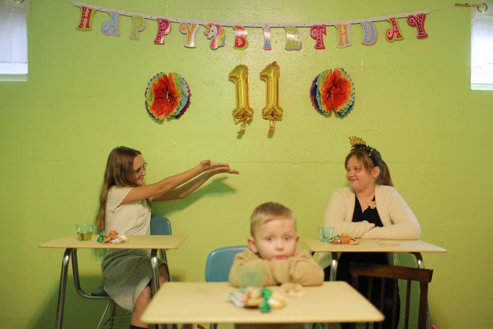 A hanging sign reads Happy Birthday 11 over two girls interacting. A young boy is in the foreground with his head on a desk.