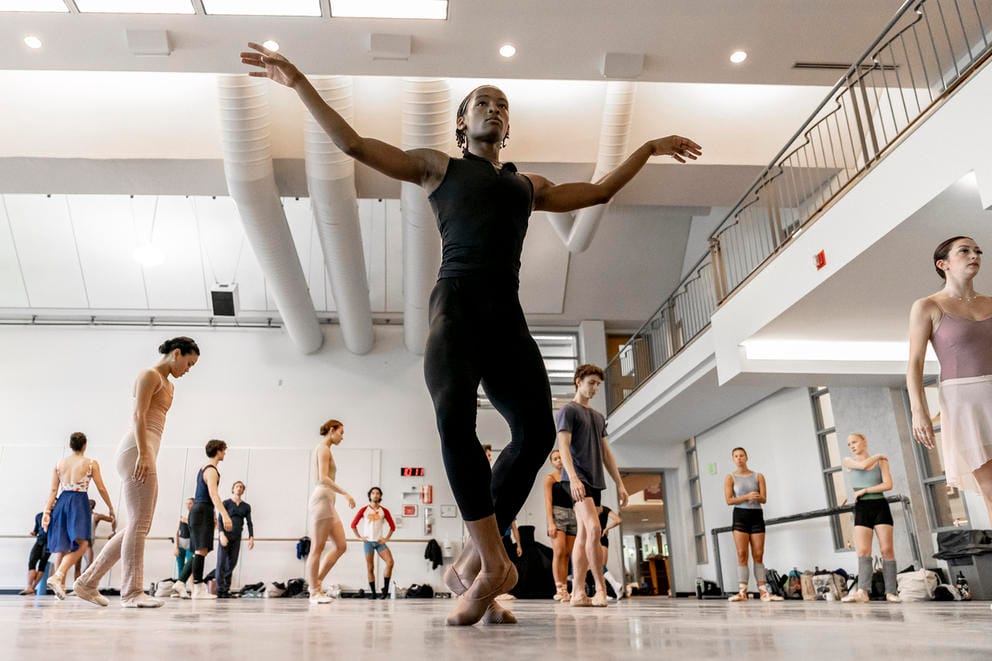 A ballerina at the front of the image outstretches his arms and plies; other dancers walk in the background