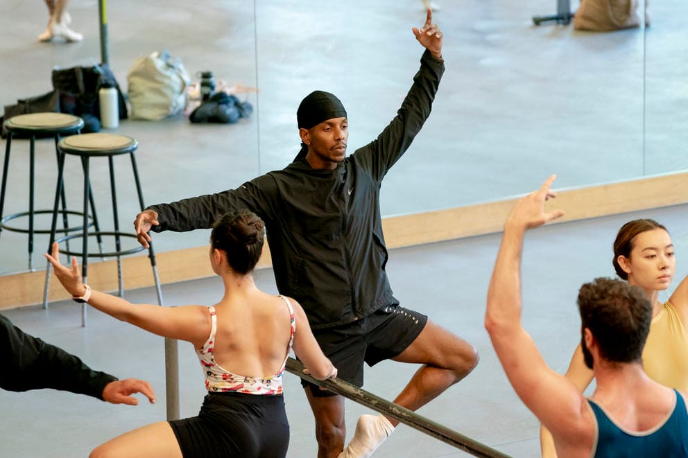 A ballerina puts one foot on his knee as he stretches at a barre; he has one arm stretched horizontally and the other outstretched above his head