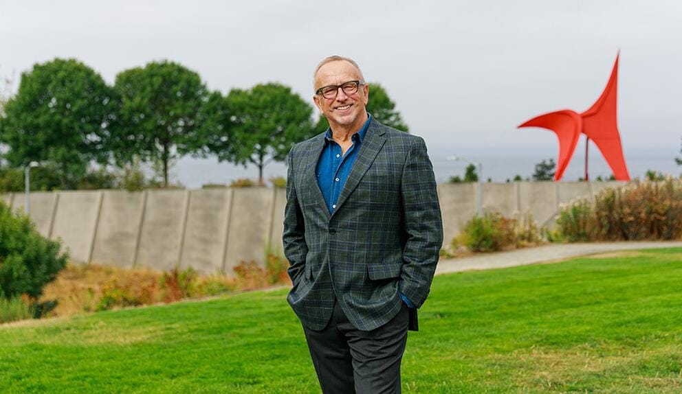 a smiling man in glasses stands outdoors in a sculpture park