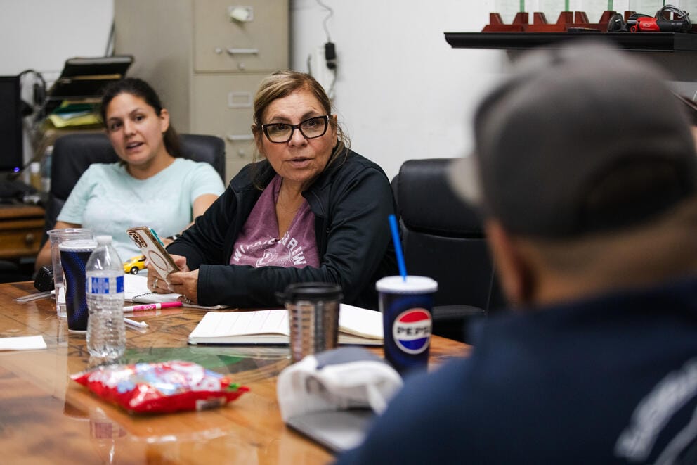 Two women, facing the camera, talk to a man with his back to the camera at a conference table in an office