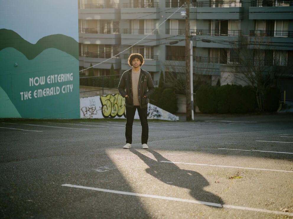 A full body shot of a man standing in a parking lot. He is backlit by the sun and his shadow is visible stretching out in front of him. The wall behind him reads