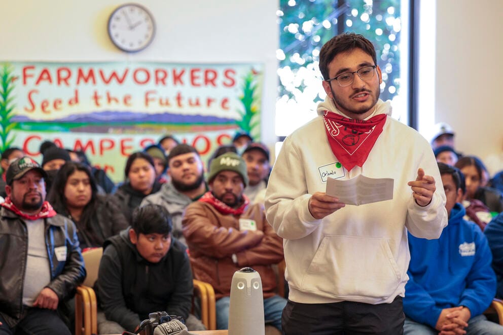 A man wearing a red handkerchief offers testimony in a crowded room.