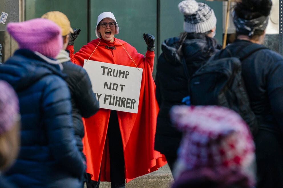 A woman dressed in a red cloak with a sign reading 