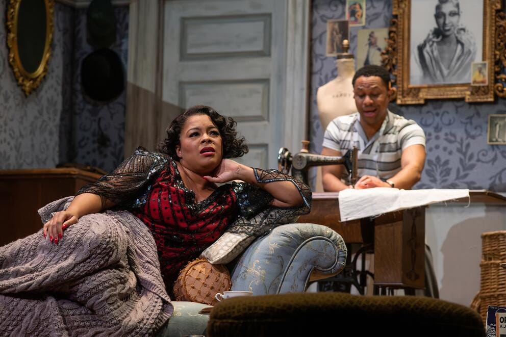 photo of a stage play in which a woman and man are seated in a 1930s living room