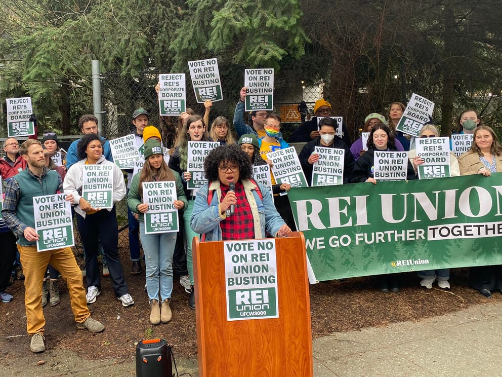A person at a podium speaks as a large group of people holding a sign that says 